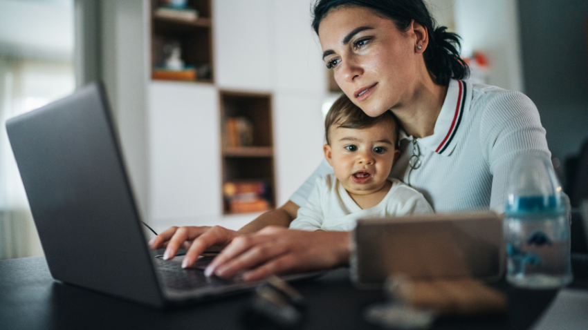 A woman with dark hair works on a laptop at a table, with a baby leaning against her arm, looking towards the viewer. A smartphone and baby bottle are also on the table. The background is a blurred home interior.