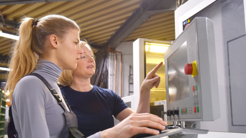 Two women are working in what appears to be a factory or workshop setting, looking at a machine control panel. A younger woman with blonde hair in a ponytail, wearing a light grey turtleneck and overalls, is operating a mouse. An older woman with blonde hair, wearing a dark blue t-shirt, is standing next to her, pointing at the screen. The screen displays various buttons and what looks like industrial controls, including a prominent red emergency stop button.